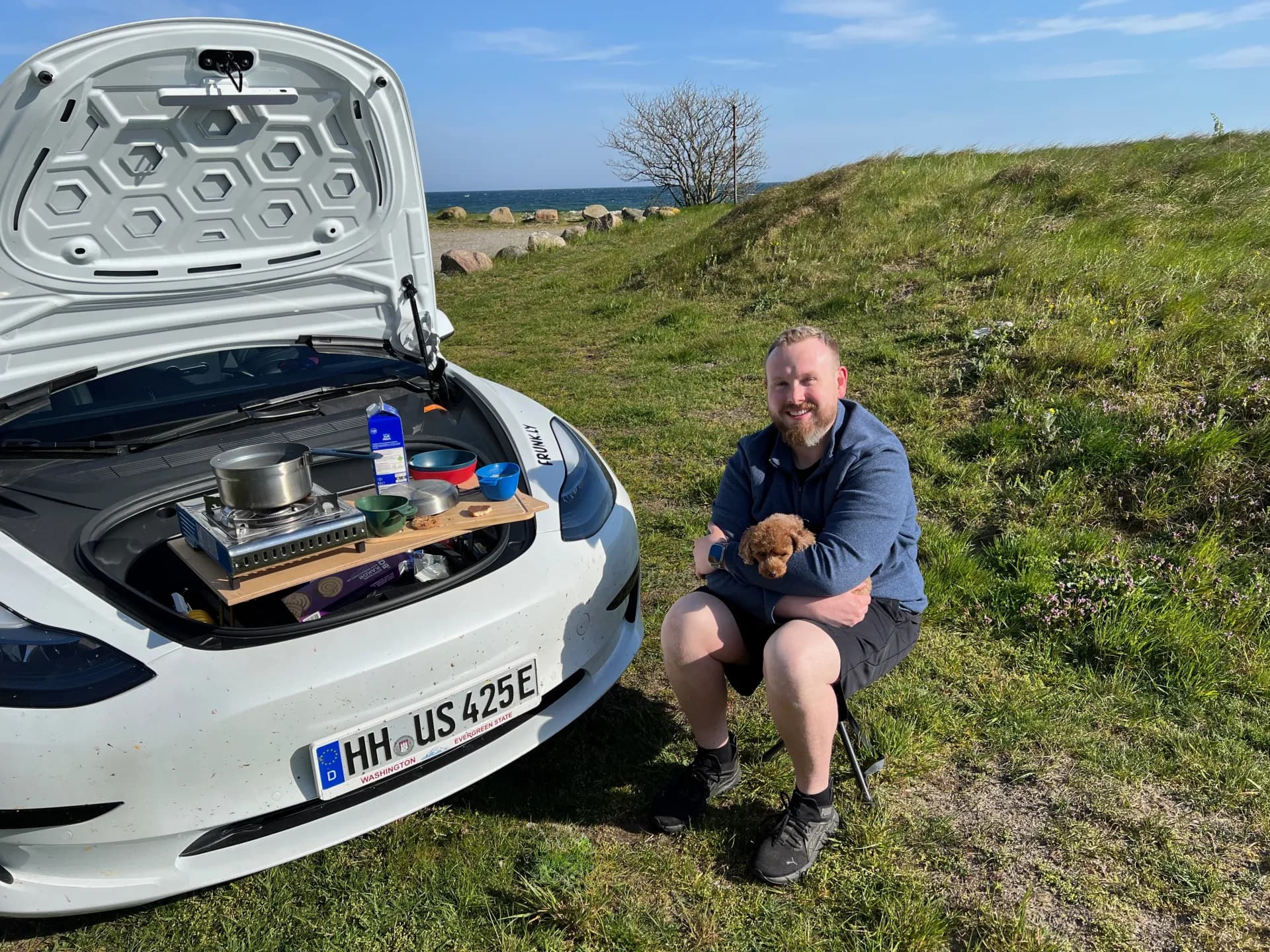 Man with dog next to Tesla Model 3 with Frunkly table cooking setup at coastal seaside spot